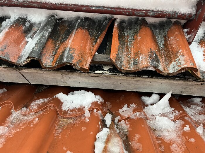 Tegelröda takpannor med snötäcke och trasig del, indikerar möjlig vattenskada och behov av reparation.