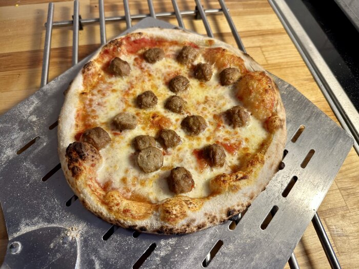 Nybakad pizza med köttbullar på metallspade och träbänk.