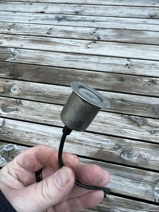 Hand håller ett litet metallbägare mot träplankgolv utomhus. Enkelt, ålderdomligt, verktyg eller redskap.