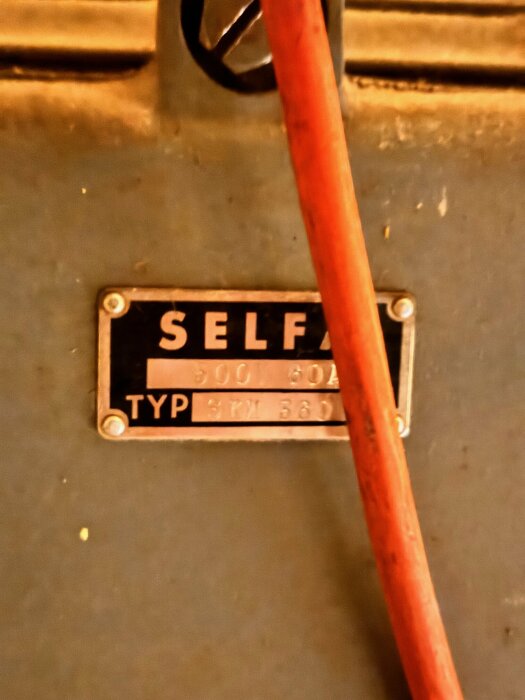 Delvis oskarp bild med röd stång, svart namnskylt "SELF" på maskindel, texten "900 60A TYPE 54TW 365".