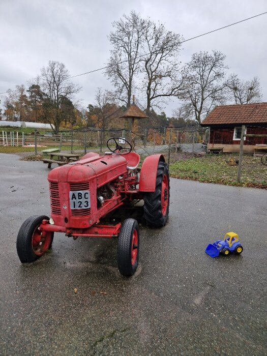 Gammal röd traktor, leksakstraktor, våt asfalt, träd, molnig himmel, gårdsbakgrund.
