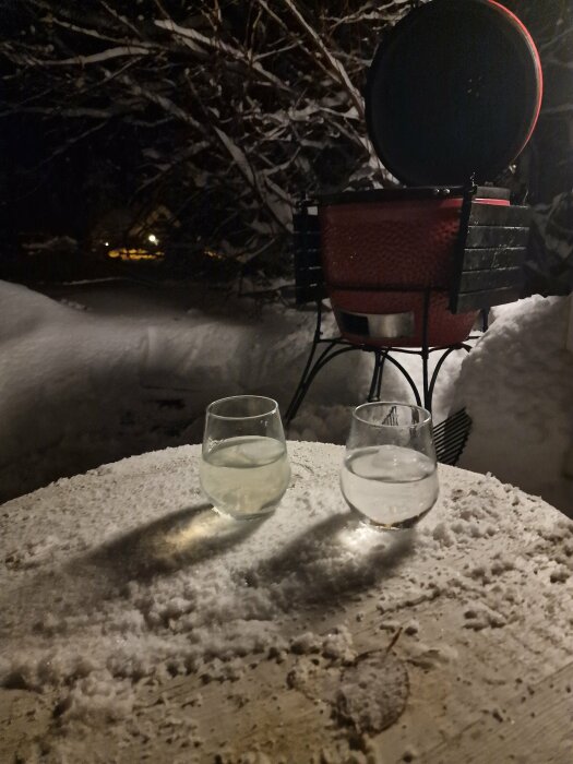 Två glas på snötäckt bord, utevärmare, mörk vinterkväll, träd i bakgrunden, mysig atmosfär.