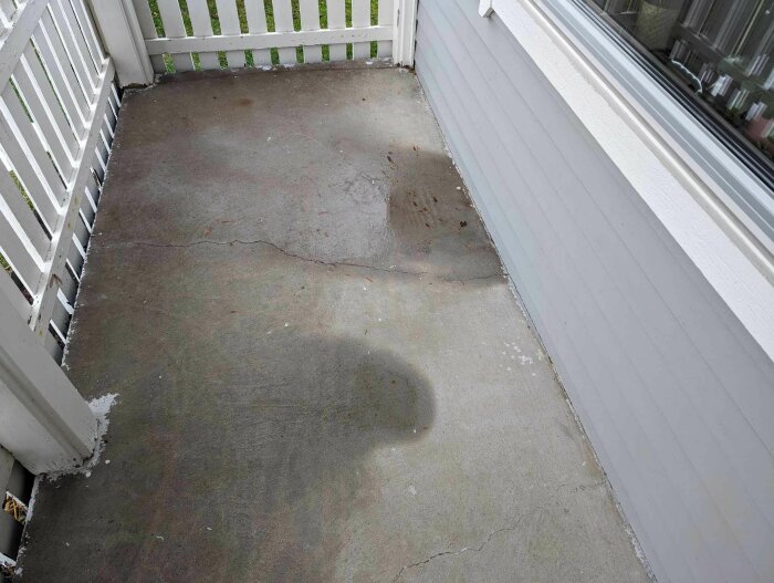 En veranda med trästaket, ett fönster, och en våt fläck på betonggolvet.