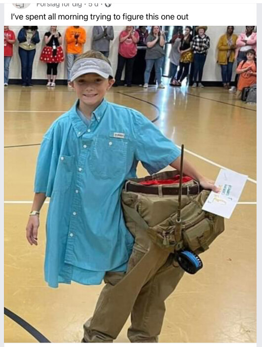 Barn i stor skjorta och byxor håller upp illusion av en kort kropp, omgiven av publik inomhus.