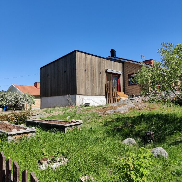 Modern trähus, trädgårdsland, klart väder, grönskande vegetation, blå himmel, ingen person synlig.