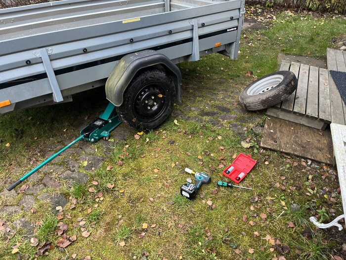 Släpvagn med lyftdäck, reservdäck på marken, domkraft, verktyg och batteridriven skruvdragare.