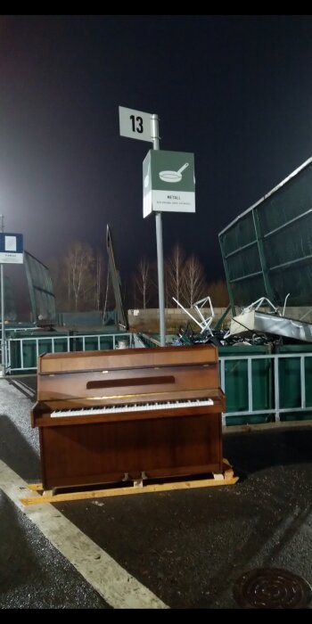 Övergivet brunt piano vid återvinningsstation, platsnummer 13, mörk kväll, container för metallavfall, sopor i bakgrunden.