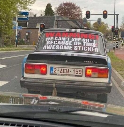 Bakre delen av en bil med humoristisk klistermärkestext om begränsad sikt, stannad vid trafikljus.