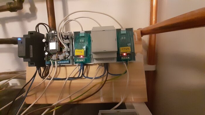 Elskåp med automationsutrustning, kopplingar, kablar och LED-indikatorer monterade på träplatta, omgiven av rörinstallation.