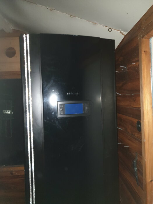 Svart kylskåp med blank yta framför träpanelvägg, inomhus, svagt belyst, ser nött ut.
