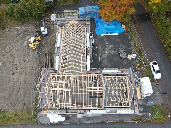 Flygbild av en byggarbetsplats med halvbyggd husstomme, byggmaterial, fordon och höstträd runtomkring.