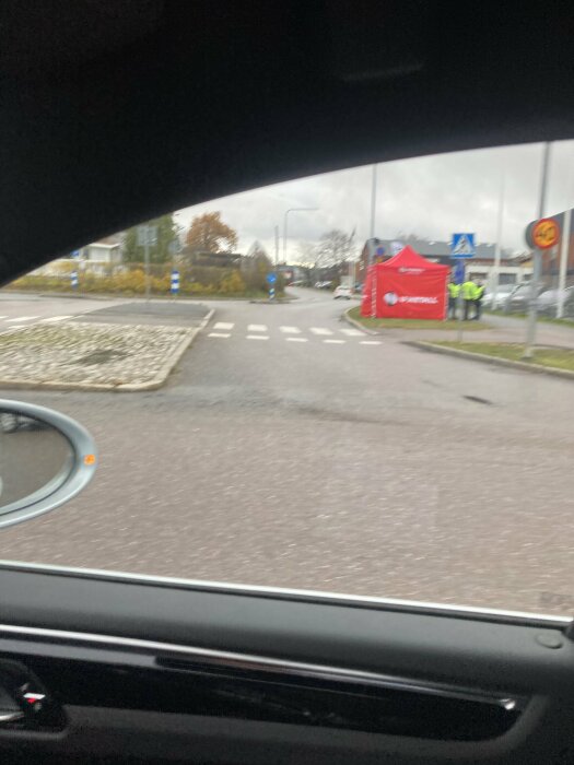 Bild tagen från en bil visar en gata, röd reklampaviljong, och personer i reflexvästar.