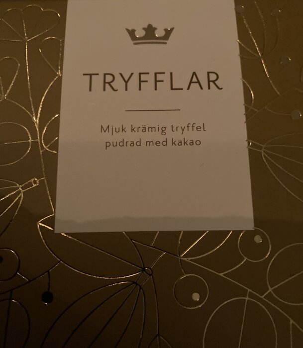 Etikett med text "TRYFFLAR", beskrivning av en mjuk, krämig tryffel pudrad med kakao, mot dekorerad bakgrund.