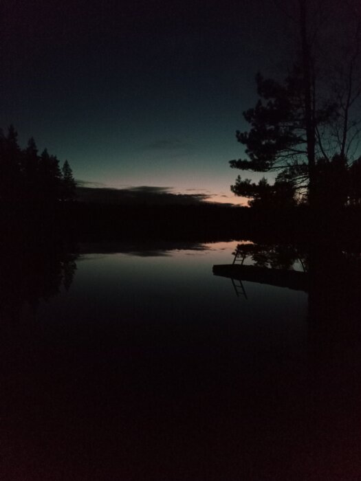Kvällshimmel speglar sig i lugnt vatten, silhuetter av träd, avlägsen brygga, skymningens stillestånd.