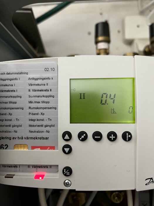 Elektronisk termostat med display, rör i bakgrunden, knappar, etiketter på svenska, röd indikatorlampa lyser.