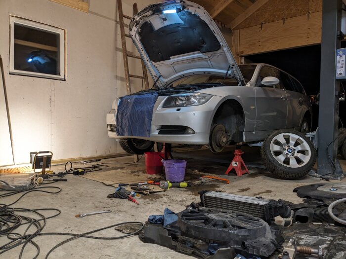 Bil på lyft i garage, öppen motorhuv, verktyg och bildelar på golvet. Reparation eller underhåll pågår.