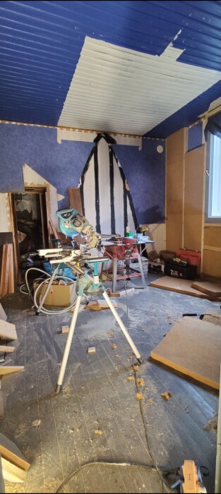 Ett rörigt rum under renovering med blått tak, verktyg, spån, och byggmaterial.