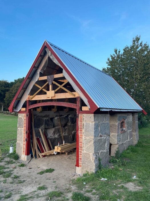 En delvis byggd eller renoverad stenlada med röda detaljer och blå himmel i bakgrunden.