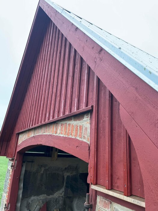 Röd träbyggnad, takkant, buad port, tegeldetaljer, öppen dörr, stenar synliga inne, strukturdelar synliga.