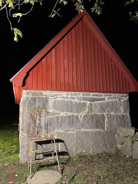 Rött trähusgavel mot nattbakgrund, stenfot, gammal fällbar stol framför, gröna löv ovanför.