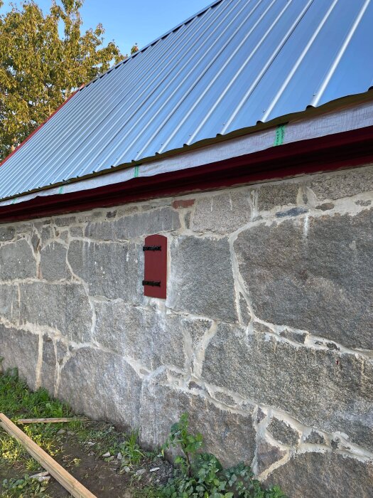 Stenhus med röd detalj, plåttak, trä och gröna växter under en klar himmel.