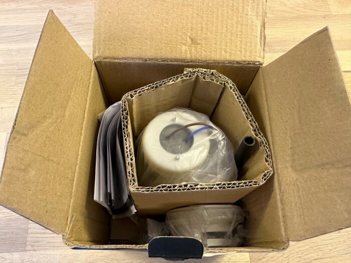 Kartonglåda öppen med packmaterial, svart objekt, hoprullade pappersark, plastomsluten vit rund sak.