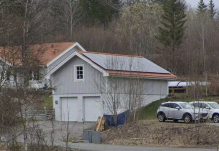 Ett vitt hus med garage, bilar framför, träd i bakgrunden och mulet väder.