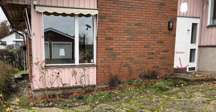 Delvis murat hus med tegel och rosa panel, fönster, dörr märkt "Bastun", trädgårdsväg, yttre markeringar runt kabel.