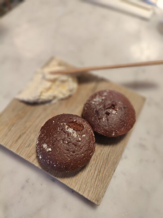 Två chokladmuffins på träbräda, florsocker, smörkniv och smör på köksbänk.