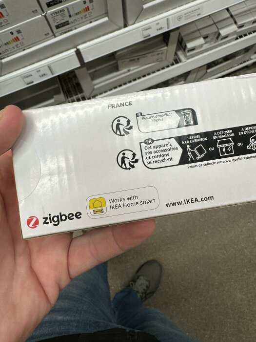 Produktförpackning med Zigbee-logotyp, IKEA Home Smart-kompatibilitet, återvinningsinstruktioner, person i affär.