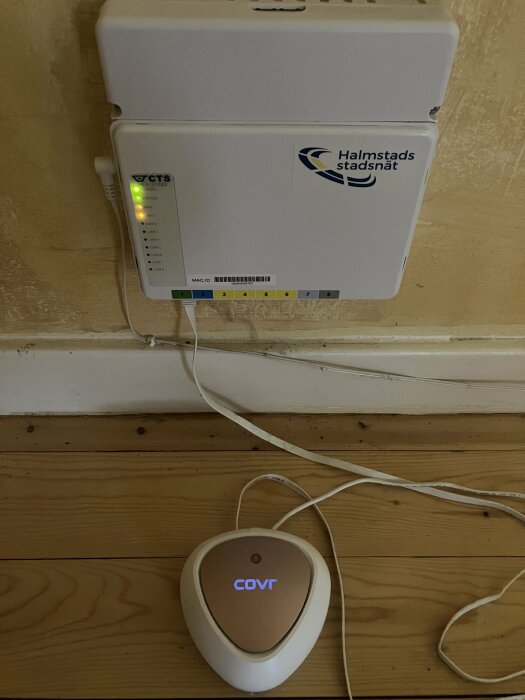 Fiberanslutningsbox från Halmstads Stadsnät och en WiFi-router med märkningen 'COVR'. Sladdar och trägolv syns.