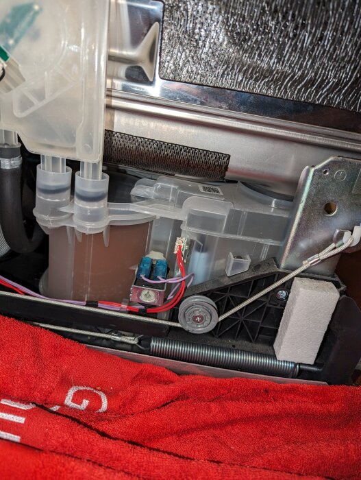 Interiör av maskin, möjligen tvättmaskin, med elektroniska komponenter, pumpar, sladdar och en röd trasa.