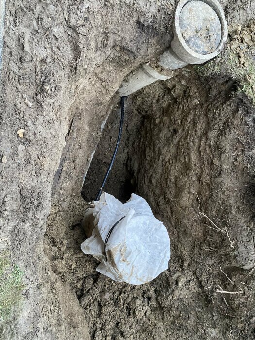Ett rör sticker ut ur marken nära en grävd grop med en påse eller presenning i botten.