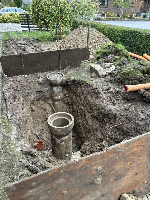 Grävd grop med avloppsrör, betongbrunnar i trädgård, pågående markarbeten, grus och jordhögar.
