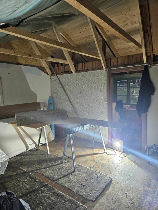 Renoveringsprojekt i oisolerat boningsrum med synlig takstol, fönster och arbetslampa som lyser.