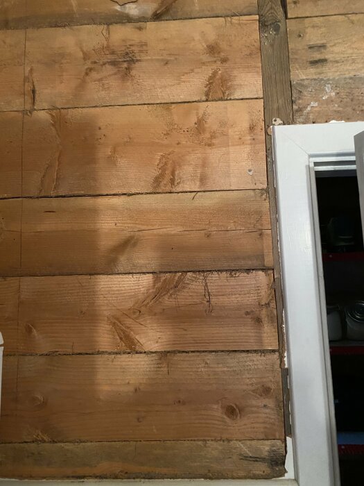 Trägolv med åldrade plattor, en vit dörrkarm till höger, delvis synligt mörkt utrymme.