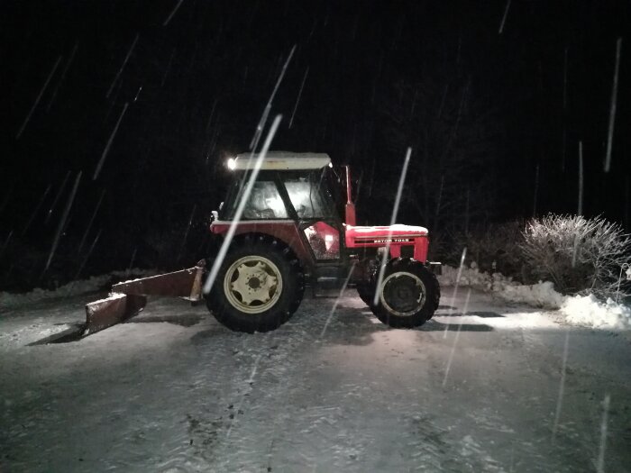 Röd traktor med snöblad nattetid under snöfall, upplyst av strålkastare, vinterarbete.