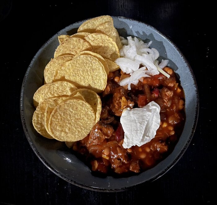 Chiligryta med bönor, lök, gräddfil och majschips på en tallrik mot mörk bakgrund.