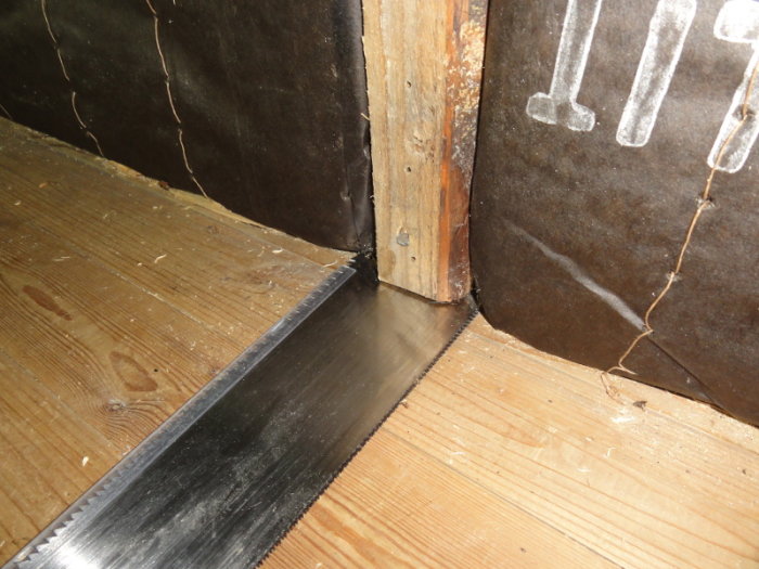 A saw blade inserted under a wall stud to assess construction; unfinished wood floor and dark wall insulation visible.