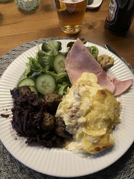 Middagstallrik med potatisgratäng, köttbullar, rödkål, skinka, sallad, öl och senap på ett bord.