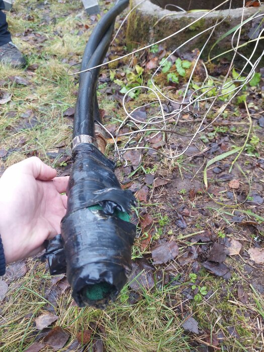 Hand håller svart tejp-lindad kabel utomhus i dagsljus, grön mark, löv och sten i bakgrunden.