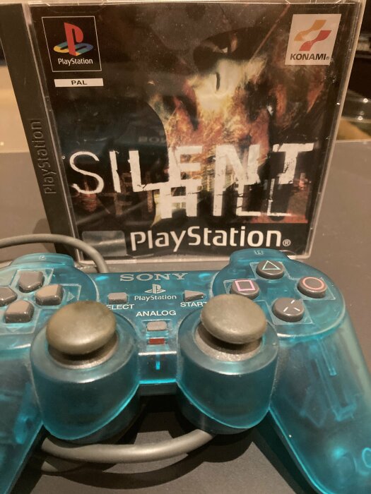 PlayStation-spel "Silent Hill" med transparent blå handkontroll framför. Retro gaming-tema, Konami, PAL-version.