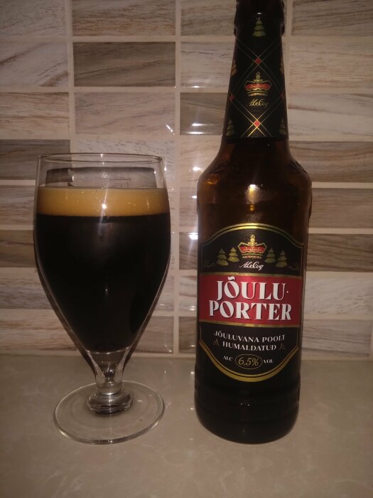 En flaska mörk öl märkt "Joulu Porter" bredvid ett glas med öl, placerade mot kakelvägg och ljus bänkskiva.