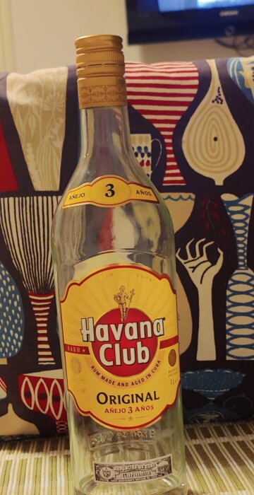 En nästan tom flaska Havana Club rom framför ett färgglatt kuddfodral, stående på ett bord.