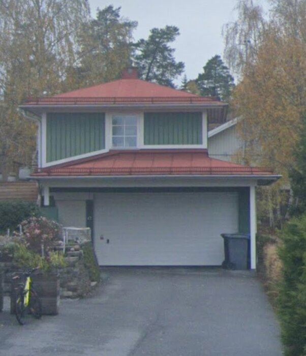 Ett tvåvåningshus med rött tegeltak, vit fasad, garage, cykel, soptunna och vegetation. Cloudy day.