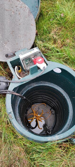 Öppen gatubrunn med elektrisk utrustning och kablar, över brunn med vatten och alger. Gräs runtomkring.