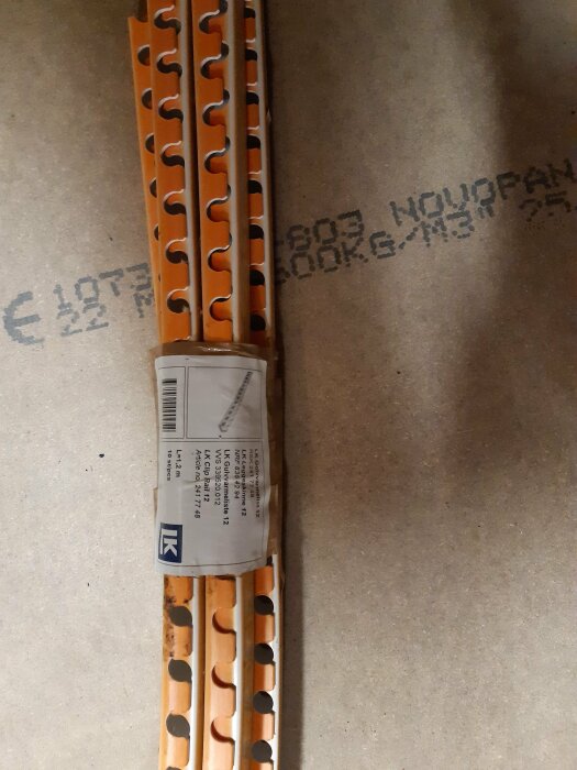 Förpackade orange och svarta plastdelar på betonggolv, med streckkod och fraktsedel, sannolikt byggmaterial.