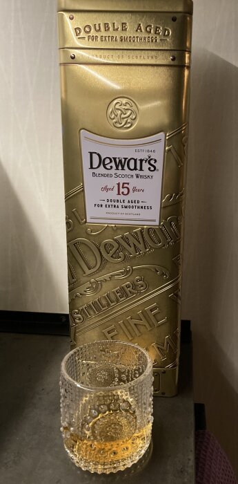 Guldig whiskyförpackning märkt "Dewar's", glas med whisky, "Aged 15 Years", skotsk, dubbelåldrad för släthet.