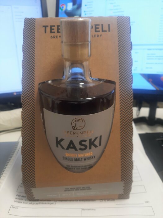 En flaska single malt whisky märkt "KASKI", sherry lagrad, framför en datorskärm, står på papper.