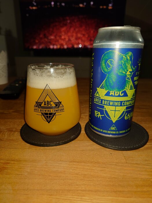 Ölglas med skum på underlägg, Apex Brewing Company IPA-burk, TV i bakgrunden.